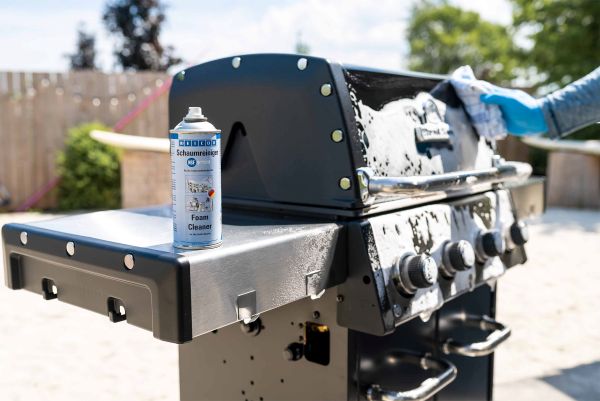 A can of WEICON Foam Cleaner rests on the side shelf of a barbecue grill covered in cleaning foam while a gloved hand wipes the surface.