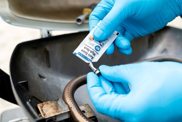 A person wearing blue gloves applies WEICON Anti-Seize High-Tech paste from a small sachet onto a metal screw near a grill component.