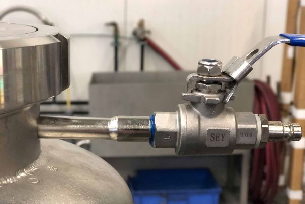 A stainless steel ball valve with a blue lever is mounted on a pipe connection of a metallic vessel.