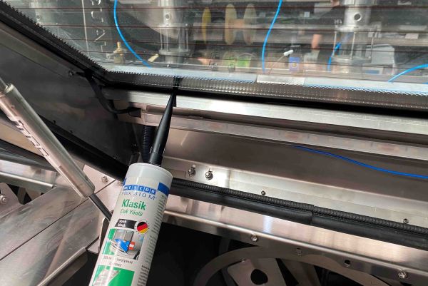 A cartridge nozzle applies WEICON sealant along the inner frame edge of a glass cover on an industrial machine.